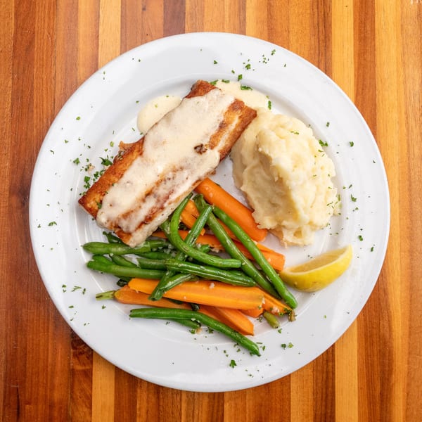 Pan-seared fish with creamy sauce, mashed potatoes, green beans, carrots, and a lemon wedge.