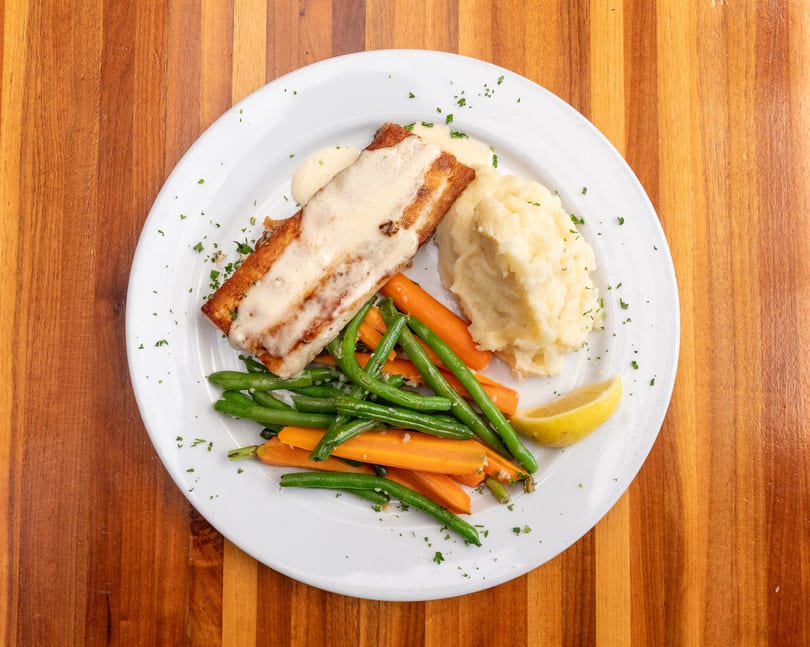Pan-seared fish with creamy sauce, mashed potatoes, green beans, carrots, and a lemon wedge.