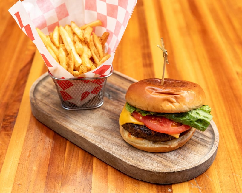 Cheeseburger with lettuce and tomato, alongside a basket of french fries on a wooden board.