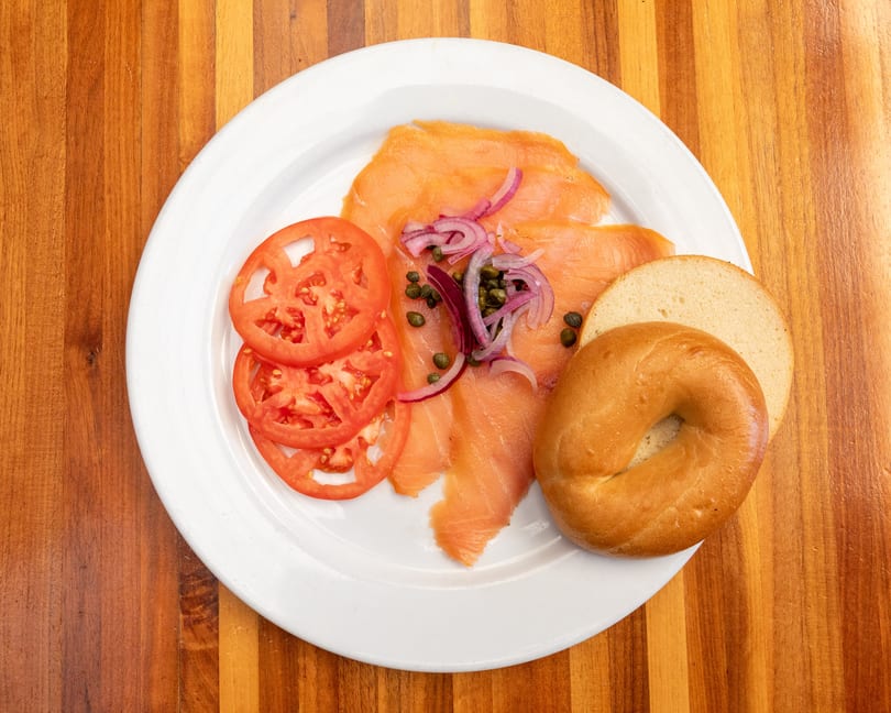Smoked salmon with capers, red onions, tomato slices, and a bagel on a white plate.