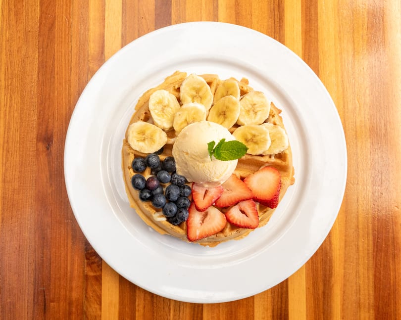 Golden waffle topped with sliced bananas, blueberries, strawberries, and a scoop of vanilla ice cream.