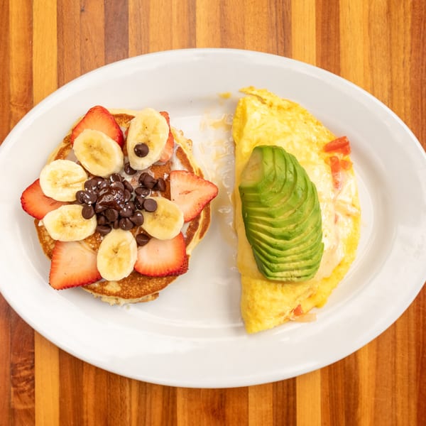 A breakfast plate with pancakes topped with fruit and chocolate chips, and an omelet with avocado.