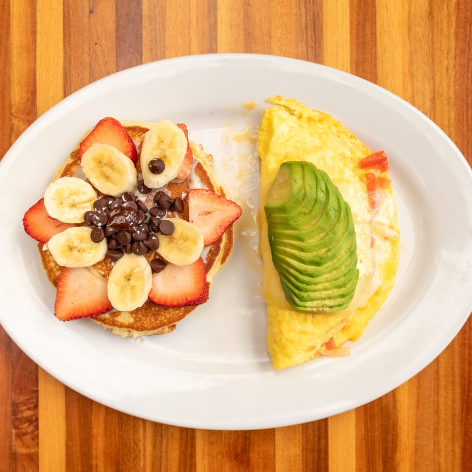 A breakfast plate with pancakes topped with fruit and chocolate chips, and an omelet with avocado.