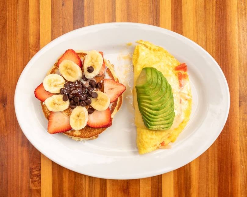 A breakfast plate with pancakes topped with fruit and chocolate chips, and an omelet with avocado.
